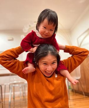 Nicole Wee, wearing a Honeycomb sweatshirt, smiling, and holding her infant on her shoulders.