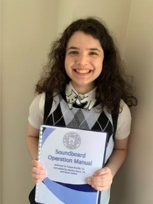 Smiling, dark-haired, high school student Cassie, wearing a gray-and-black argyle vest and a cat-motif shirt, holds a blue-and-white spiral-bound manual.