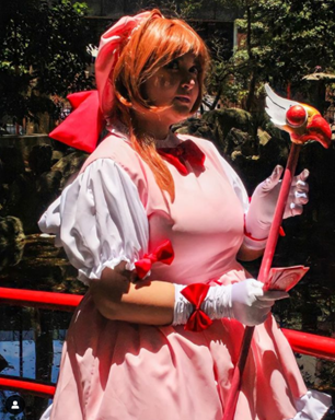 A modelo Aly Bubbly posa com o vestido rosa e branco cheio de laços cor de rosa, chapéu cor-de-rosa e a varinha mágica da personagem de anime Sakura Card Captors. Ao fundo um lago cheio de árvores ao redor.