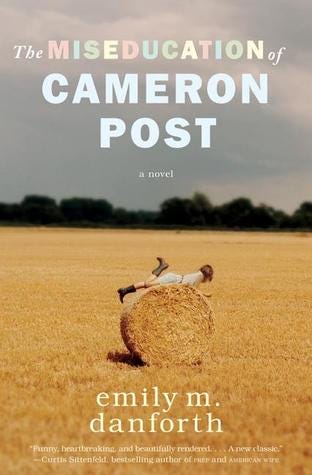 The cover of popular lesbian novel, The Miseducation of Cameron Post by Emily Danforth. On the cover of the book, a girl wearing a t-shirt, shorts and cowboy boots lays on a bale of hay in the middle of a field.