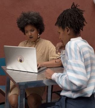 Na imagem, duas mulheres negras estão sentadas de frente para a outra e olhando para um notebook. Uma das mulheres está digitando e a outra segurando um óculos de grau com a mão perto da boca enquanto olha para o notebook.