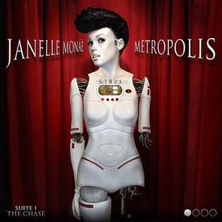 Janelle Monáe is depicted as an android with an all white mechanical looking body. Part of her left arm is missing, and she stands in front of a red curtain with the words “Janelle Monáe Metropolis” across the top.