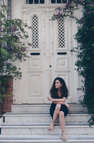 Long, dark, curly, haired slender woman sitting on the steps before a large set of white entrance doors to a home.