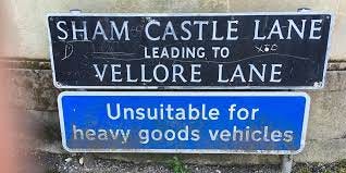 Two street signs — the top one says in white writing on a black background Sham Castle Lane leading to Vellore Lane. the second one is under it and is a bright blue background with white writing and it says: Unsuitable for heavy goods vehicles.