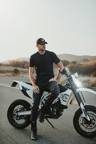 Man in black jeans, black t shirt, black boots, black cap, sitting on a motorcycle looking cool.
