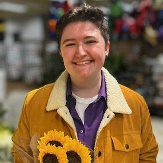 Alannah in an orange-yellow jacket, smiling, with short brown hair and a handful of daisies.