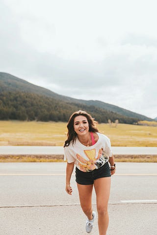 A smiling, young girl, against the backdrop of a broad road, in front of high mountains