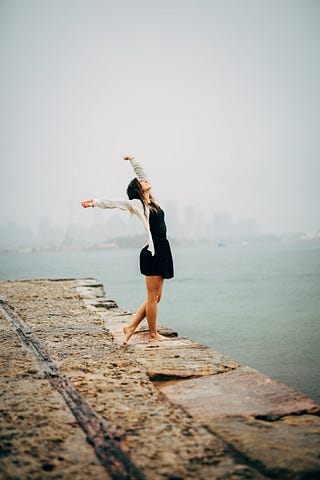 A woman stands at the edge of a stone pier. It is raining and her face is lifted to the sky. She is smiling, barefoot, and wearing a black dress. Her arms are spread high embracing the sky.