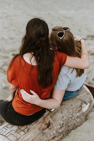 Two women sitting on a log on a beach, with their arms holding one another.