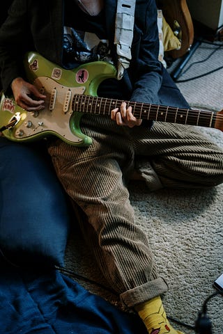 A man sits on a cushion holding a green electric guitar