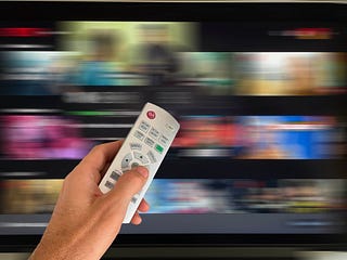 A hand aims a remote control at a screen displaying rows of key art, blurred as if they are scrolling past very quickly.