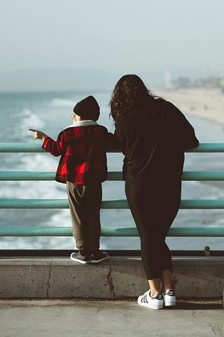 young boy standing next to his mother, overlooking the sea from a railing, viewed from the back.