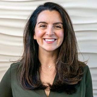 A photograph of Nicole DeCario, a woman with long brown hair smiling at the camera. She is wearing a green blouse and silver necklace.