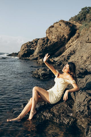 A woman sits on a rock in a white summer dress by the sea blocking the sun with her hand