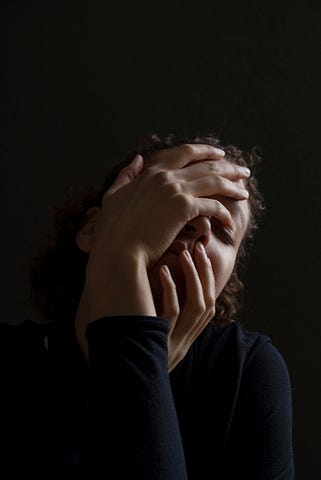 A woman in black against a black background with her hands over her face.