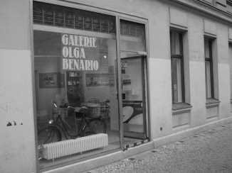 Fotografia em preto e branco da Galerie Olga Benario, em Berlim, em que o nome do estabelecimento está bem em evidência num grande vidro, que parece com uma vitrine e está na entrada do lugar.