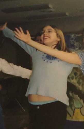 A white female child of around nine holds her arms out in a kind of dance. Another girl’s arm is in here way. She is wearing glasses and a blue shirt which has ridden up slightly so that a small amount of her stomach is visible. She is smiling.
