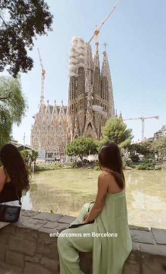 The picture shows the influencers travelling in meditterranes and especially their stopover in Barcelona. The influencer poses in green dress back in front of the Sagrada Familia, a famous church, the tones are warm because the period is summer.