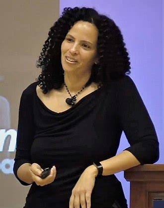A person with curly hair, wearing a black top and necklace, is standing and holding a remote control, speaking to an audience. The background includes a partial view of text on a screen or wall.