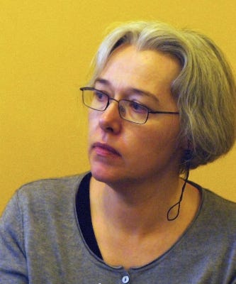 Author Susanna Clarke, a gray haired middle aged woman in this photo, wearing glasses and a gray deep v neck shirt. She appears to have an ear mic and appears to be listening to a question.