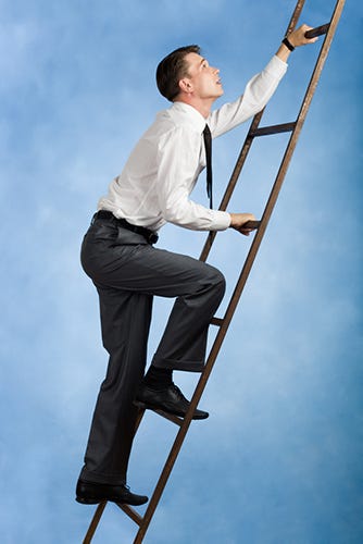 A young white man wearing black trousers, socks, and dress shoes, a white button-down shirt and tie, climbing a wooden ladder in front of a blue background.