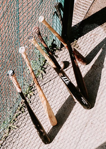 Baseball bats lined up and leaned against a green chain link fence, presumably in a dugout.