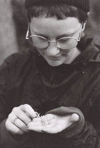 greyscale photo of Charlotte Heather, a white nonbinary person with very short hair and large round glasses. They have a serene facial expression as they look down at a small plant in their hand.