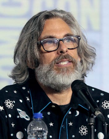 Author Michael Chabon, middle aged man with graying long hair and beard, dark rimmed glasses, and a dark blue shirt patterned with stars and planets. He’s seated before a microphone and appears to be speaking.