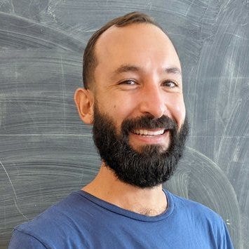 A Latino man with dark hair and a dark beard smiles while wearing a blue shirt. The man is standing in front of a chalkboard with eraser marks on the board.