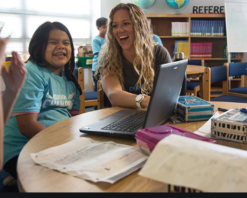 A teacher laughing and asking a kid questions while pointing at a laptop with both laughing