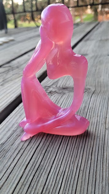 A pink resin art piece depicting a figure of a woman, one hand on her face, lost in thought.