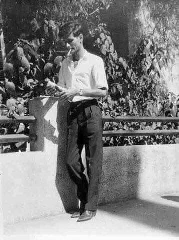 A foto é de Mahmoud Darwish parado em uma ponte. Ele está vestindo roupas e calçados pretos e brancos. O homem está parado no chão ao ar livre.