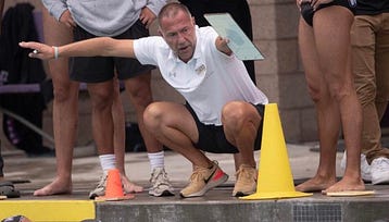 Photo of Coach Preslav Djippov crowched down in front of a yellow cone, and the edge of the pool. He is wearing a white polo shirt, black shorts, and running shoes. He is surronded by the legs of swimmers.