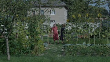 A imagem mostra Hedwig Höss e sua mãe em um jardim com elementos externos, como grama, quintal, arbustos, árvores e plantas. Ao fundo há uma casa ou prédio com janelas e paisagismo.