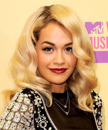 A photograph of British musician Rita Ora. She has wavy blonde hair down to her shoulders, pale skin, and red lipstick. She’s wearing a sparkly jacket over a black top. In the background, the logo of the MTV Video Music Awards is visible.