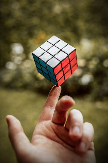 a completed rubix cube with each side a single colour, balanced on the tip of a finger against a green blurred background of grass and shrubs