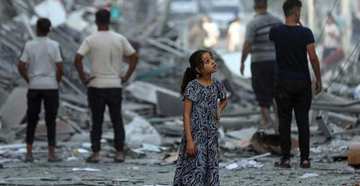 Fotografia onde mostra diversos destroços e escombros de construções palestinas, no meio da rua e destruição há uma garota palestina que olha com horror o cenário de caos e dor. No fundo, há homens palestinos que andam em meio a destruição.