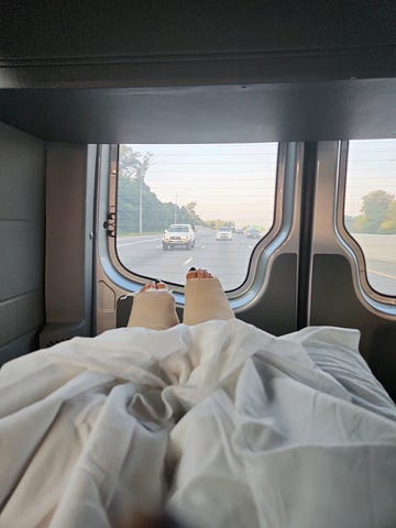 Two legs in thick casts under a blanket are seen from the inside of a hospital van. The highway stretches out beyond the back windows.