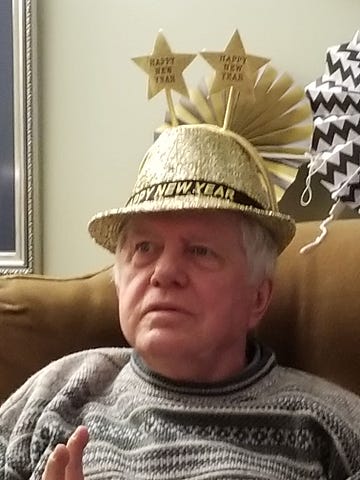 A white-haired man wearing a grey knit sweater, a gold hat that says “Happy New Year”, and a headband with two gold stars on long stalks pointing up.