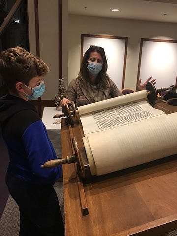 The young man reading from the Torah is Jack H. He is preparing for his Bar Mitzvah. The woman holding the Torah is Kim B., one of his teachers. The place is the chapel of Temple Shalom, where the Torah rests in the ark.