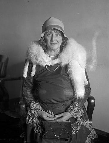 A slightly confused looking middle aged white lady sits on a chair with a white fox stole around her neck. She is Walberga “Dolly” Oesterriech. She is definitely more of a vamp than her outward appearance would suggest.