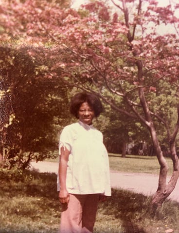 A Black woman wearing a white shirt and pink pants, standing under some trees.