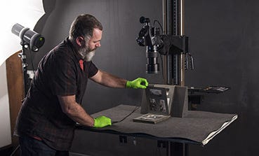 Image shows a man in a black shirt and green latex gloves using a camera on a stand to take photographs of a photo album with a lighting rig behind him.