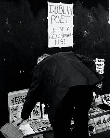 Photograph books of a poet being displeyed in astreet of Dublin, with a man picking one of the books and a sign on the wall and above the man, that says: “Dublin Poet, I‘d be a God anywhere else”.