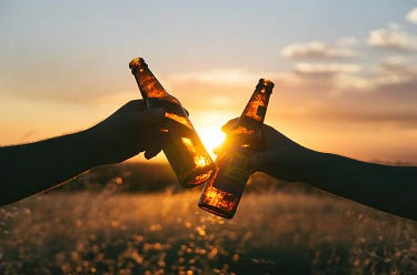 Two hands holding beer bottles clink in front of a plain with a sunset behind.