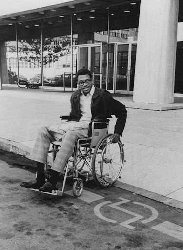 A black man uses a curb cut to descend with his wheelchair from a sidewalk to the street.