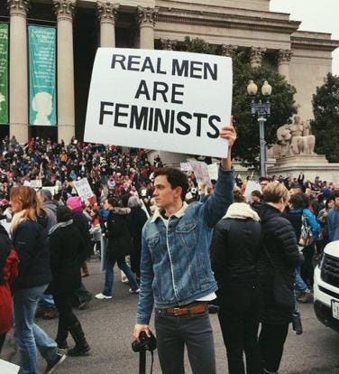 Imagem de um homem numa marcha das mulheres em que segura um cartaz que diz “Homens reais são feministas”.