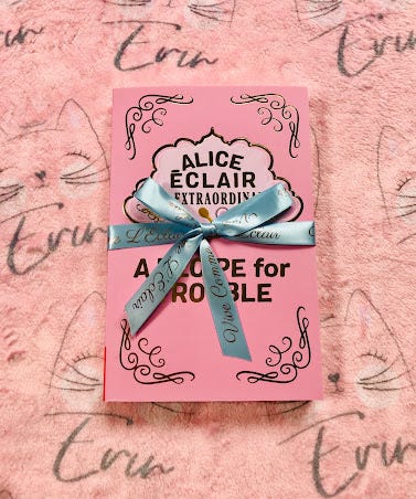 Proof copy of the book showing a less elaborate pink cover with silvery gold scrollwork in the corners and Alice’s trademark elegant title design, also in silvery gold with a pale pink background. The book has been tied with a blue ribbon by publishers. The book sits atop a pink fleece sheet with a cat logo and Erin's name emblazoned.