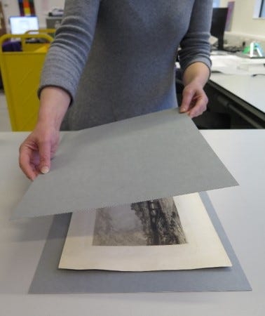 A conservator placing a slightly larger sheet of grey archival card over a black and white historic print. The print is already sitting on another slightly larger sheet of grey archival card.