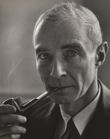 J. Robert Oppenheimer, in a black and white photo, peers into the camera while holding a pipe in his mouth. Smoke billows out from the pipe.
